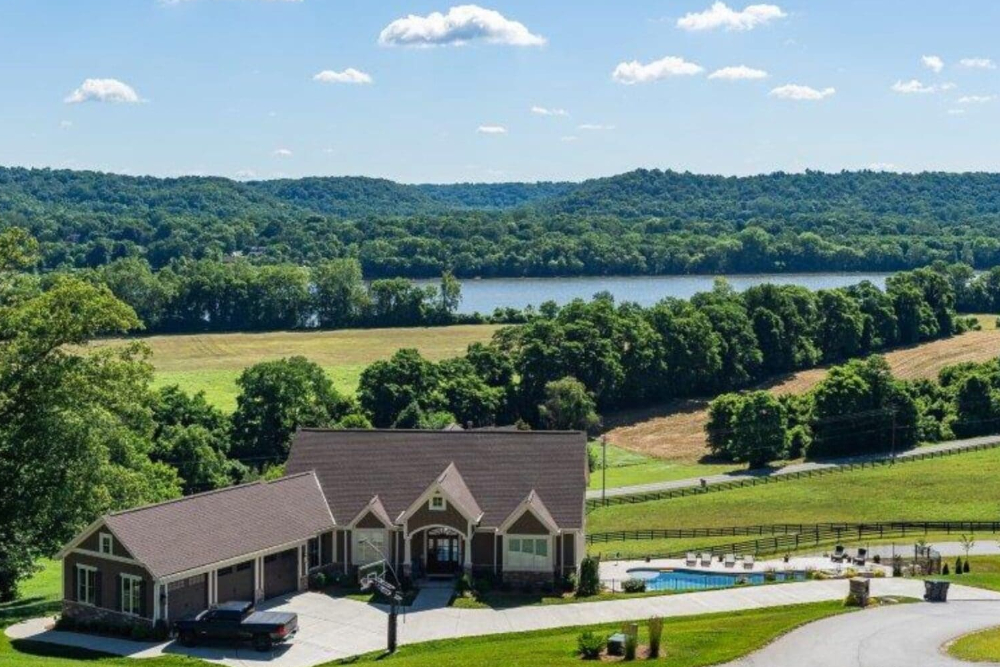 Hebron-Kentucky | Davis Landscape Design & Installation Aerial view of a ranch-style house with a brown roof and attached garage, a swimming pool to the side, surrounded by green fields and trees with a lake in the distance.